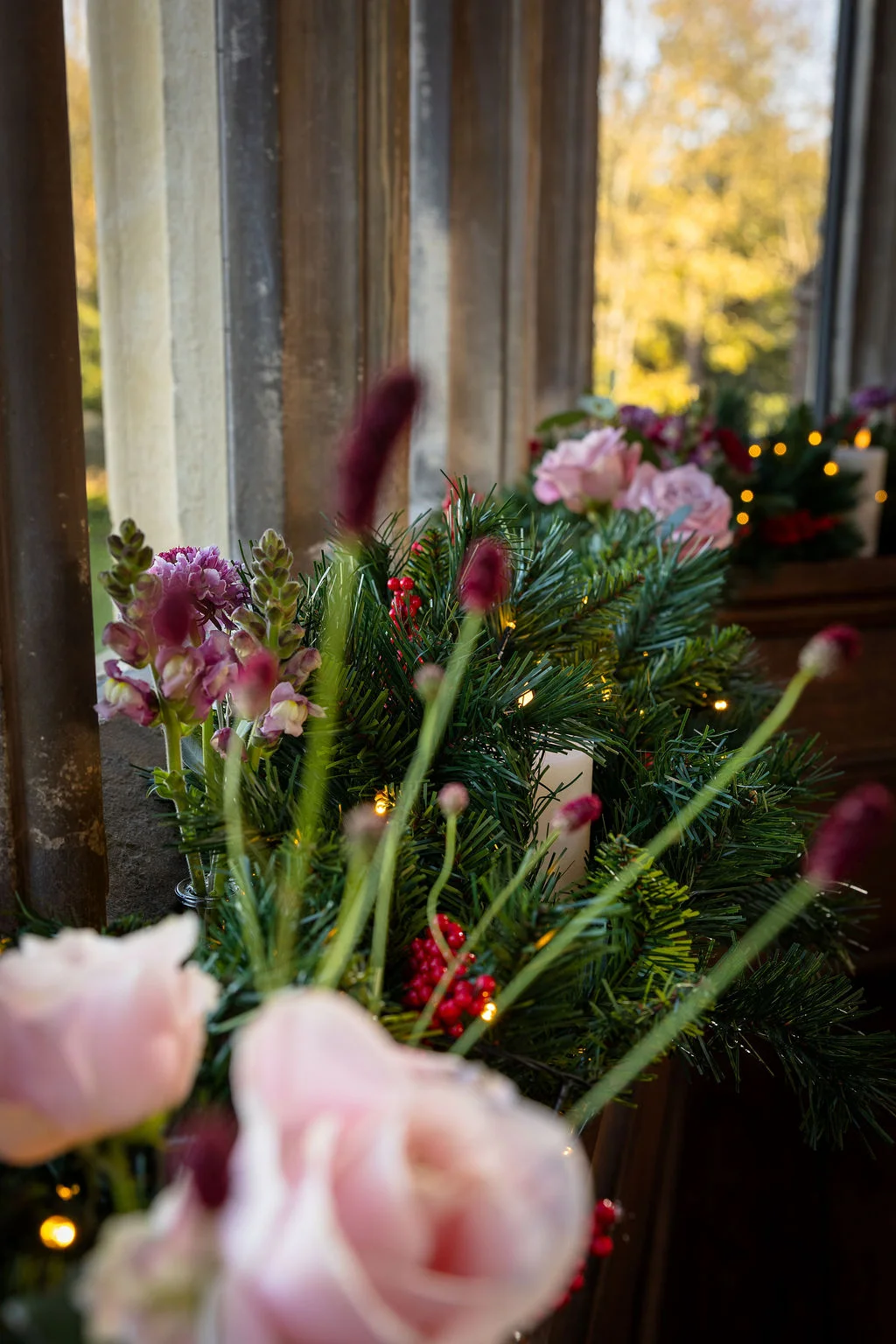 A close-up view of a stone window sill adorned with festive evergreen foliage, pink roses, delicate stems, and glowing pillar candles for a winter wedding.
