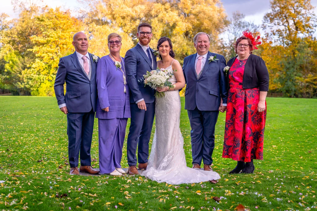 A bridal couple and family members pose for a formal group portrait on a vibrant green lawn scattered with autumn leaves, surrounded by golden trees.