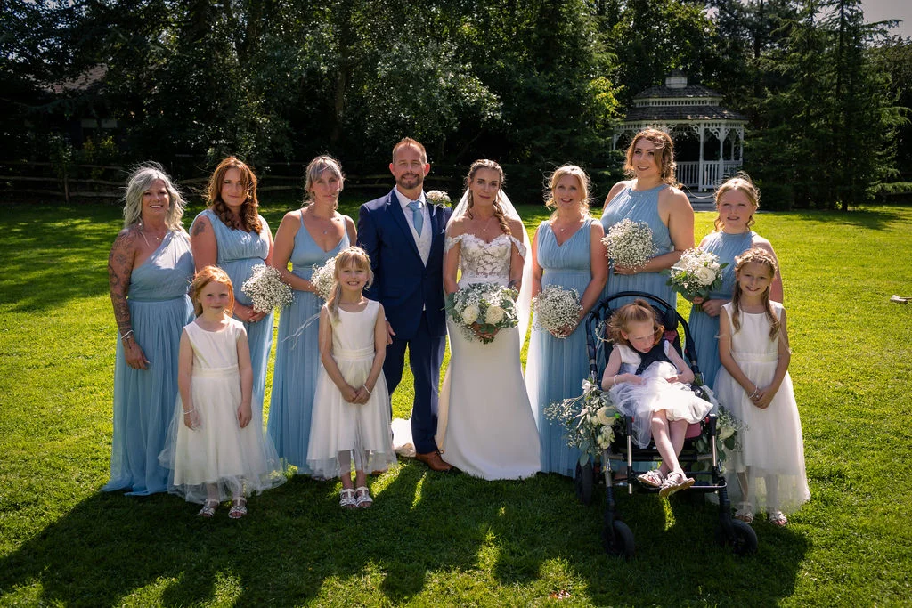 A bridal party poses outdoors on a sunlit lawn next to a garden gazebo, featuring bridesmaids in dusty blue dresses, flower girls in white, and the bride and groom at the center.