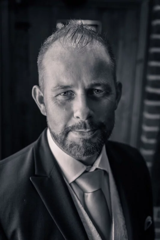 A striking black and white portrait of a groom wearing a tailored suit and tie, captured with moody window light.