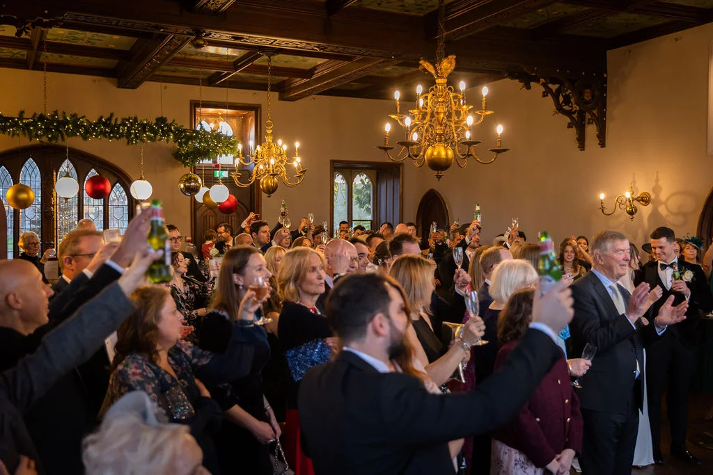 Wedding guests raise glasses and cheer enthusiastically during speeches inside a grand, wood-beamed hall illuminated by ornate golden chandeliers.