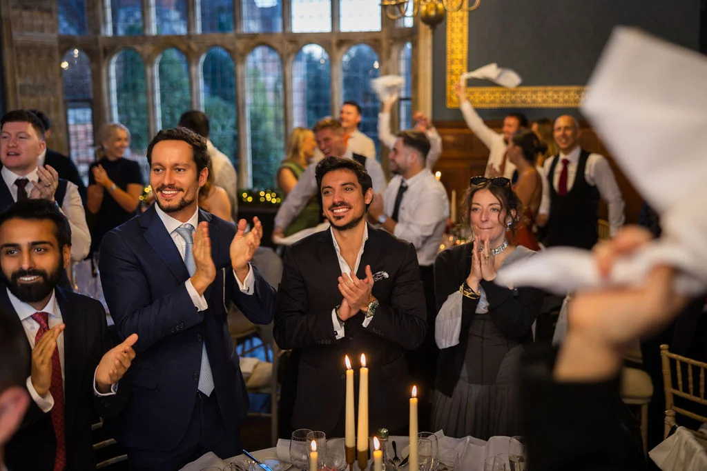 Wedding guests enthusiastically clap and cheer during speeches in a grand historic hall illuminated by candlelight.
