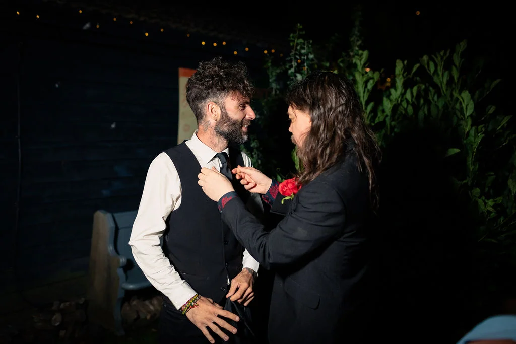 One groom gently adjusts the tie of another groom during evening wedding preparations outdoors.