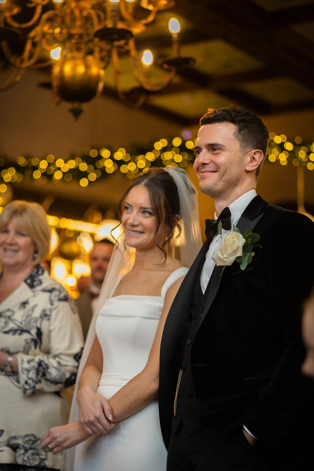 A joyful newlywed couple stands side by side during their ceremony, with the groom in a classic black tuxedo and the bride wearing an elegant white gown and veil beneath warm indoor lighting.