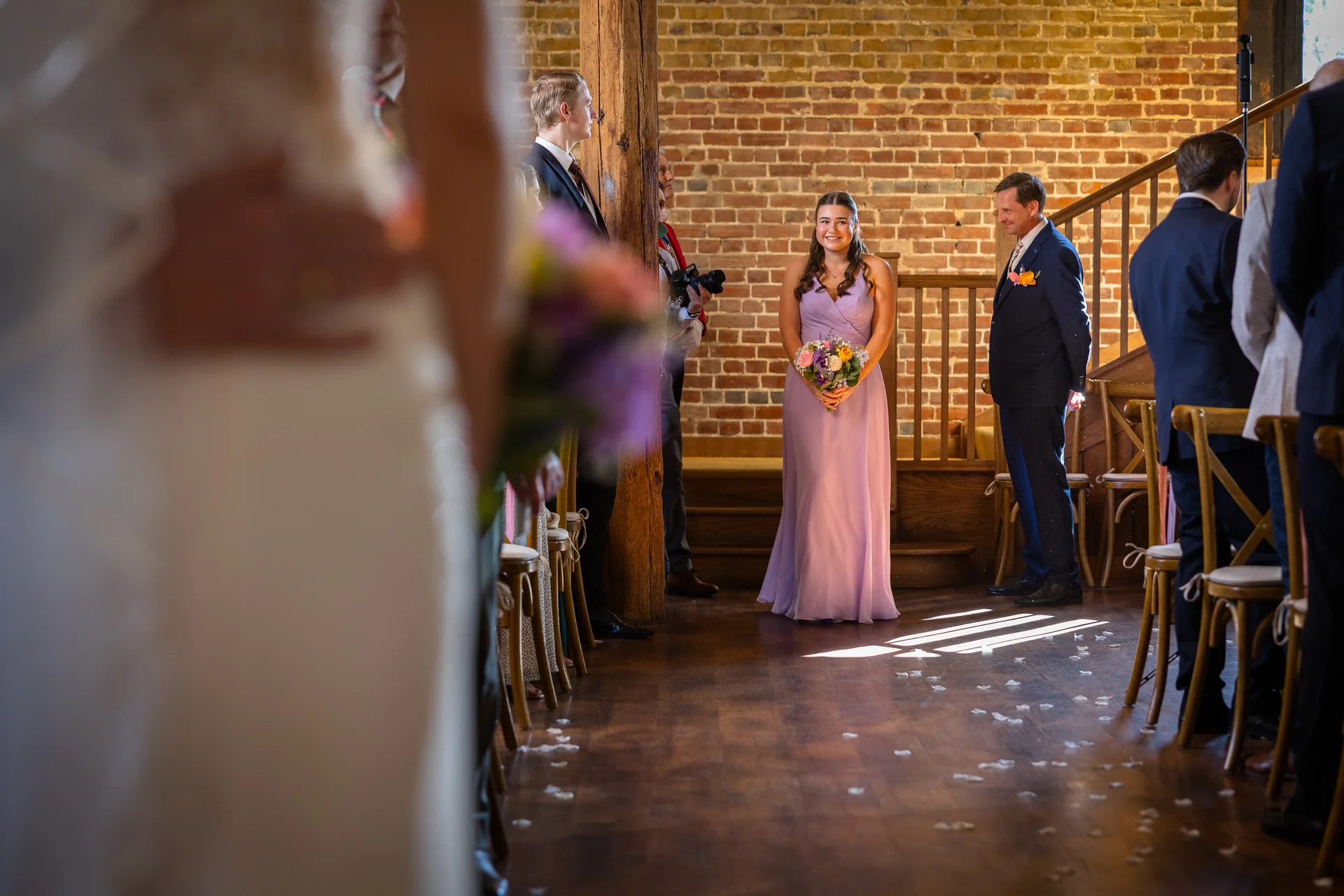 A bridesmaid in a lilac dress holds a bouquet while standing at the front of a rustic barn ceremony venue, framed by guests in the foreground.