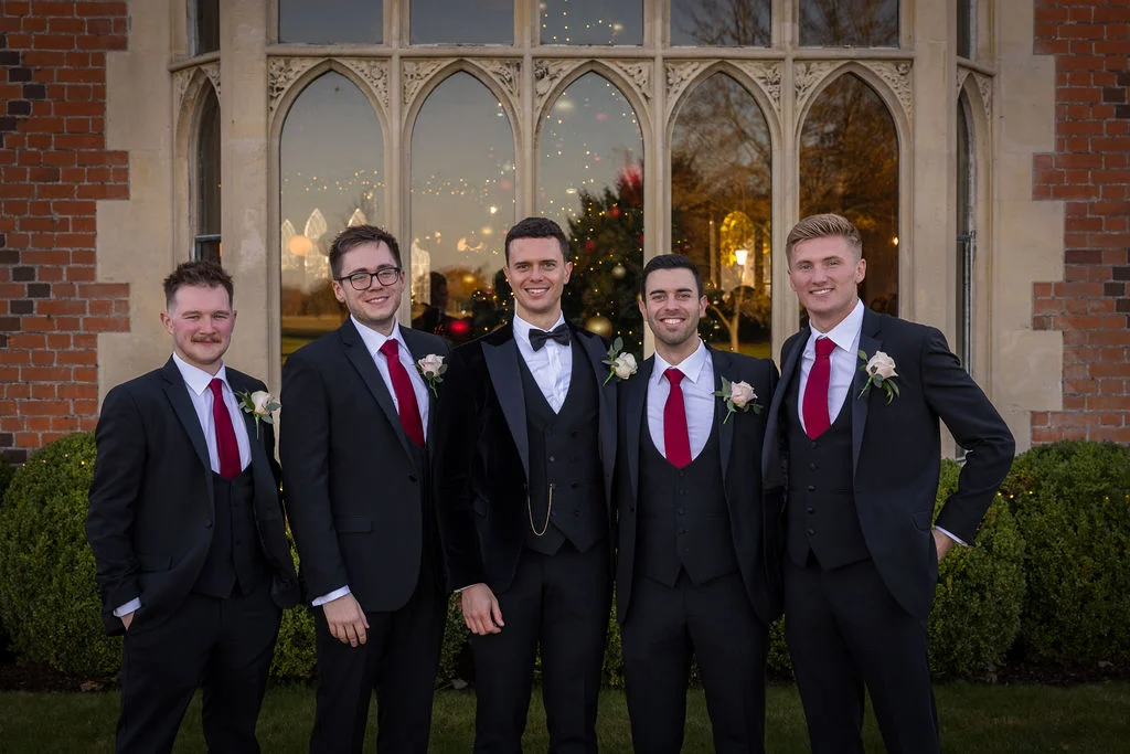 The groom and his four groomsmen pose smiling outside a grand brick manor house with large arched windows at dusk.
