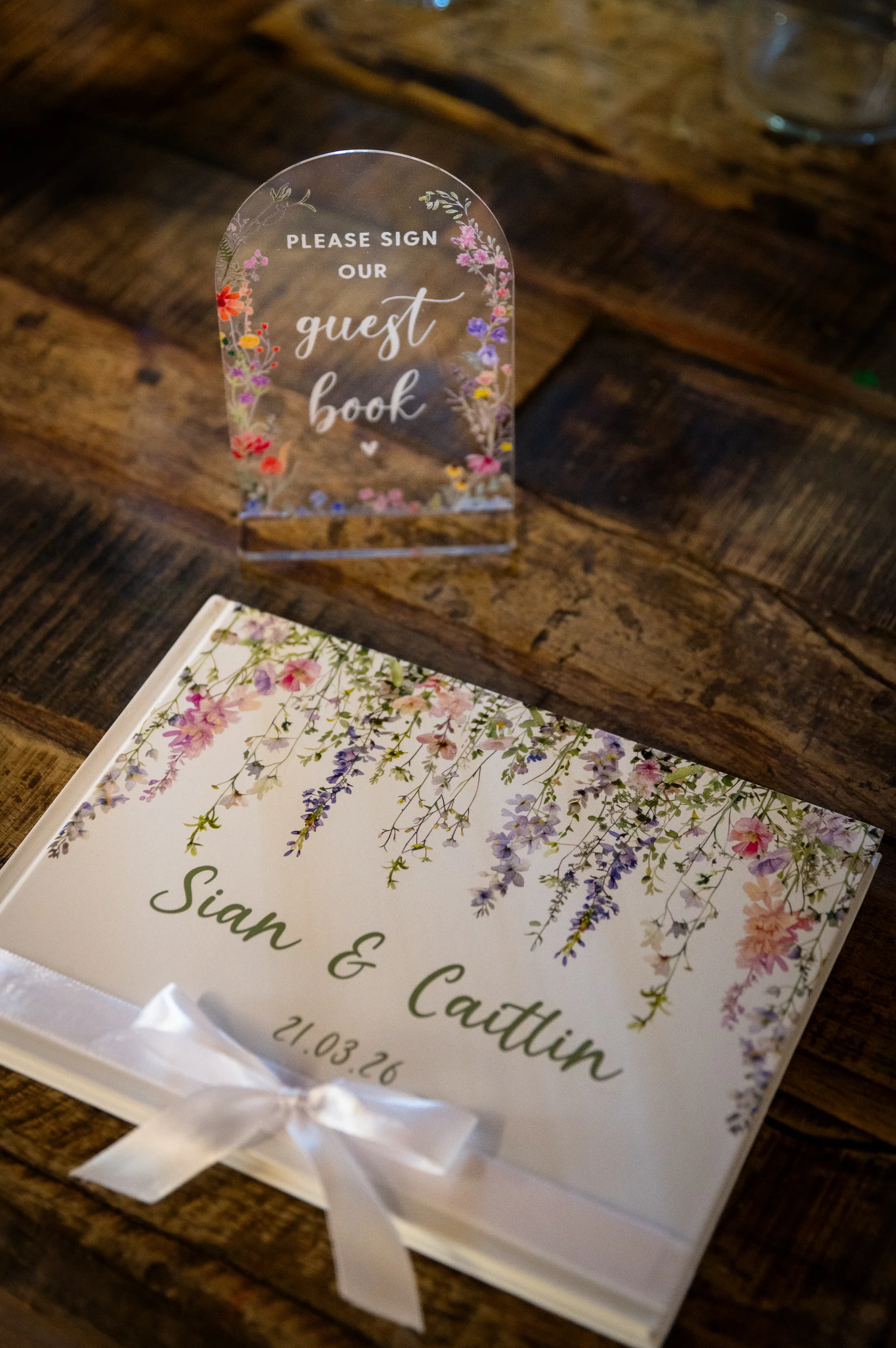 A personalised floral guest book and an arched acrylic guest book sign rest on a rustic wooden table at a wedding.