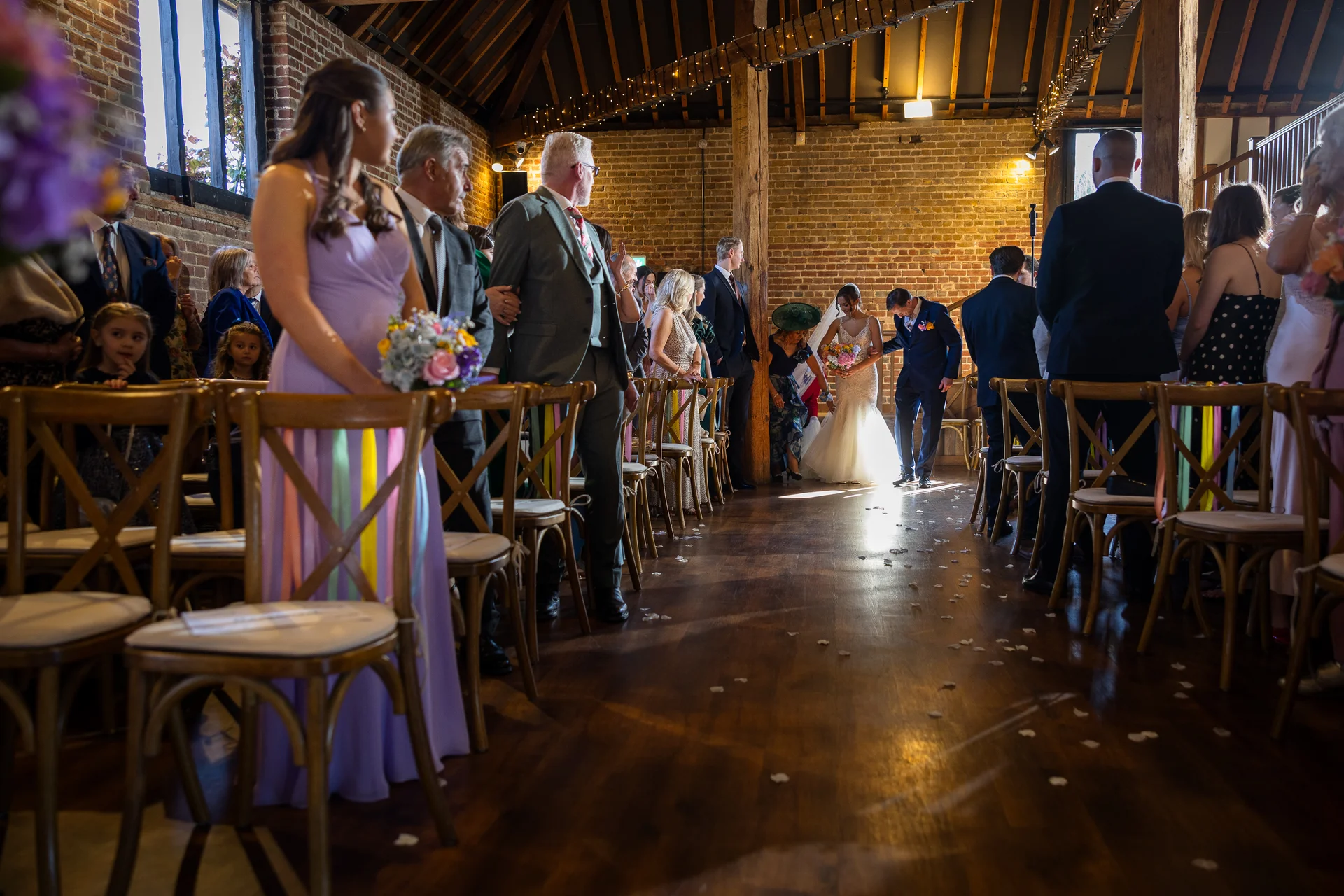 The bride and groom walk down the aisle together inside a rustic brick barn filled with seated wedding guests.