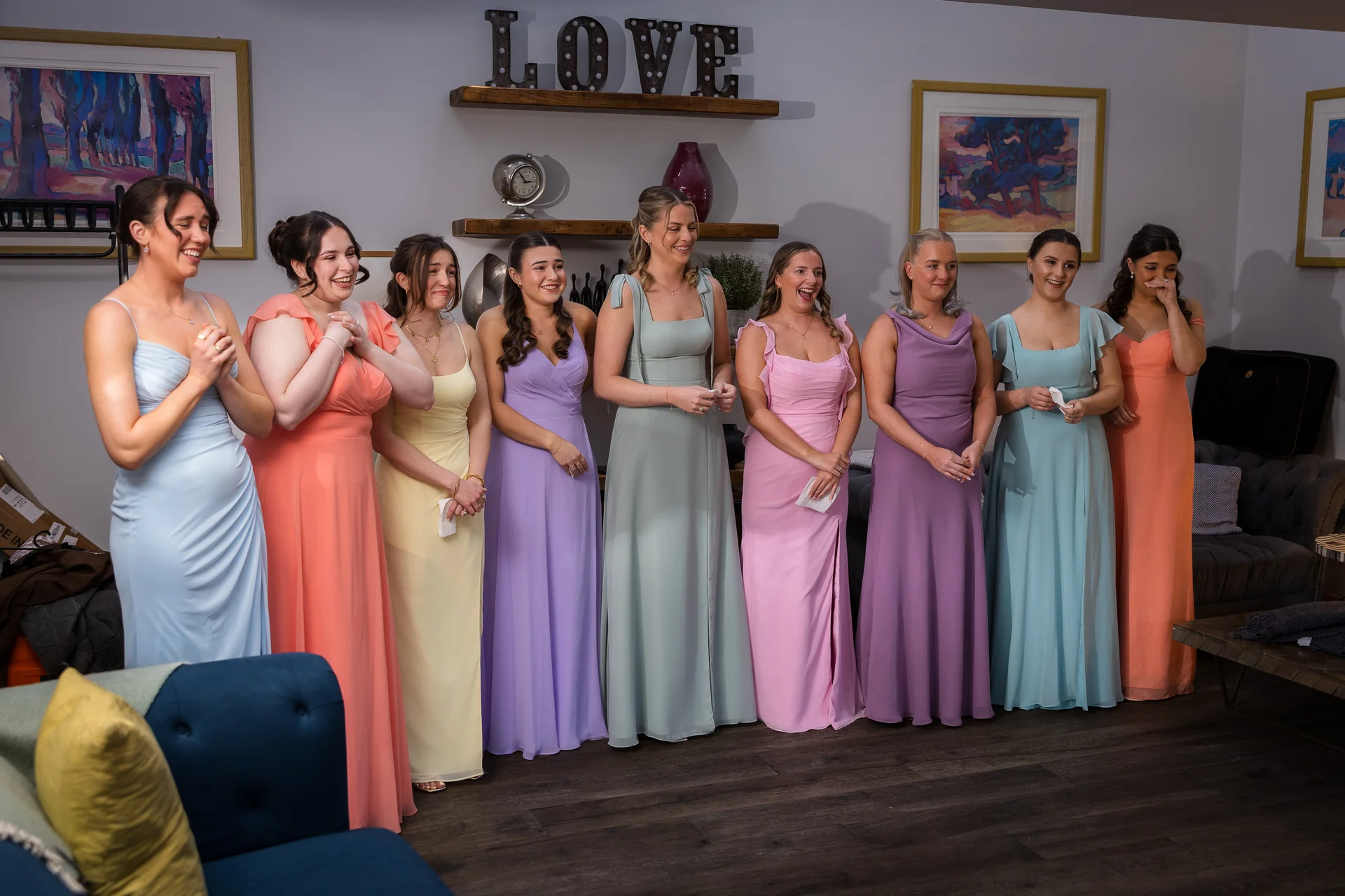 A line of bridesmaids wearing colourful pastel dresses stand together indoors, laughing and reacting with joyful emotion.