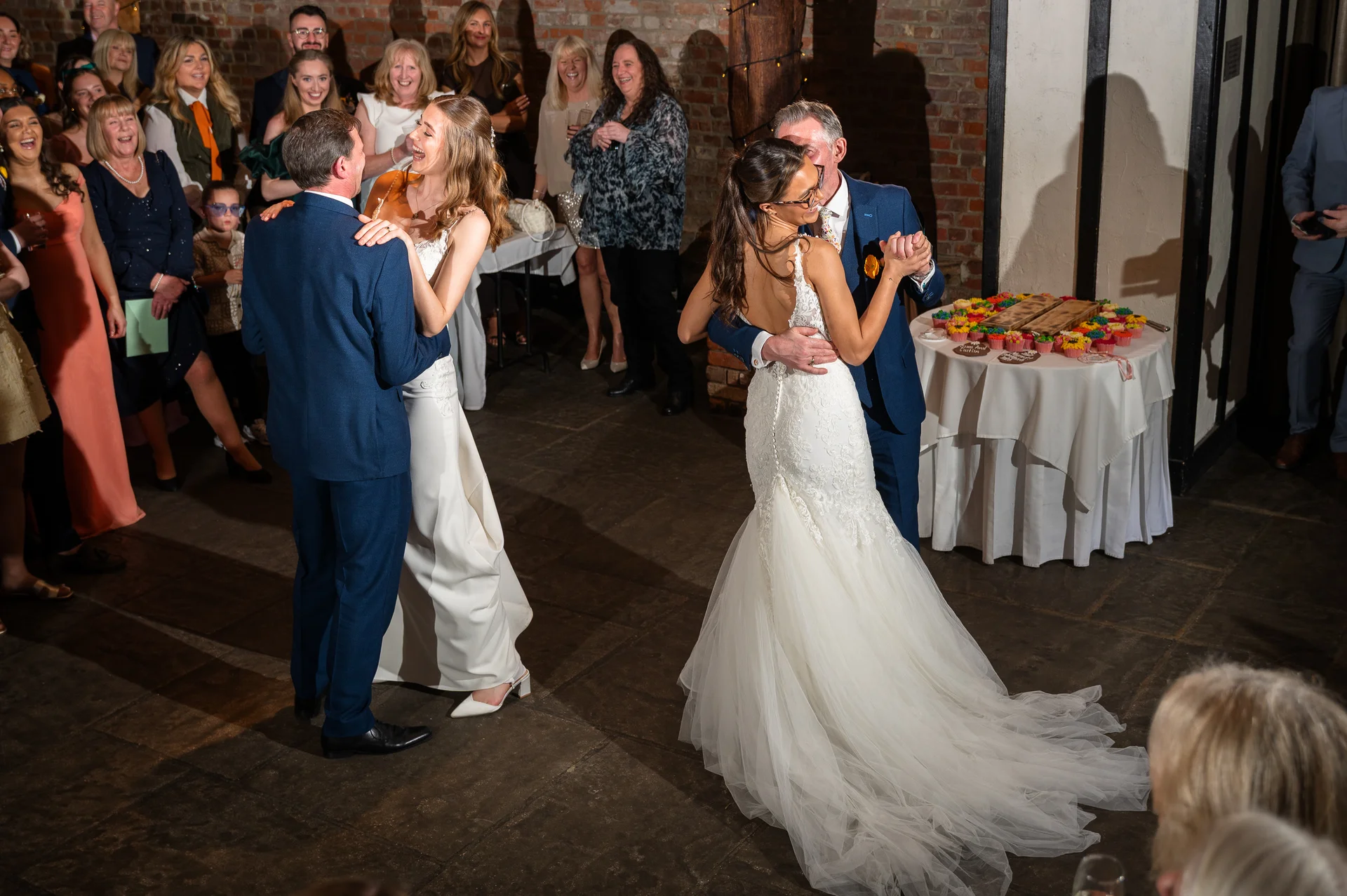 Two couples share a joyful first dance on a stone floor in an exposed brick venue while wedding guests watch and laugh.