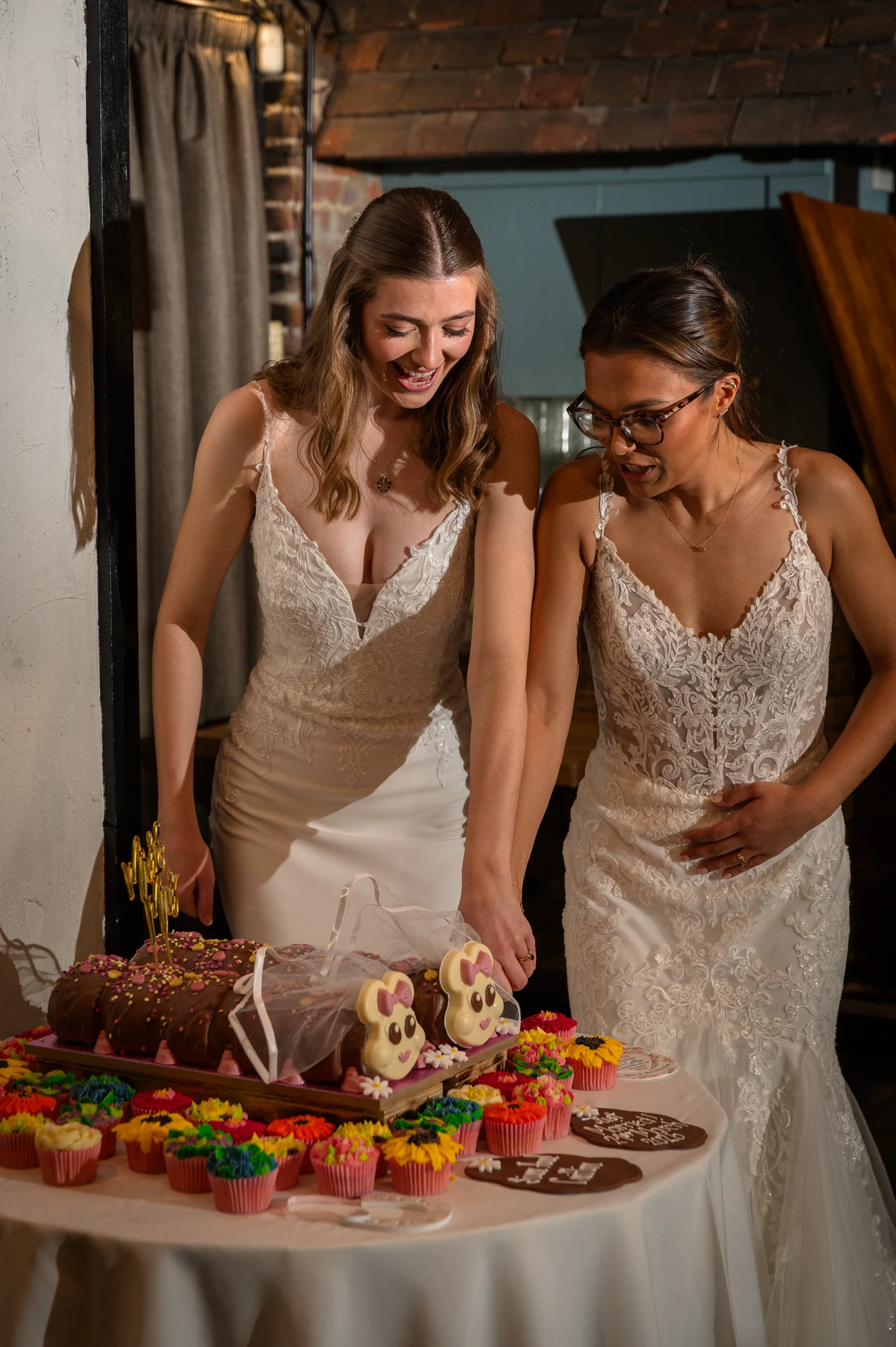 Two smiling brides in white lace wedding dresses happily cut a novelty caterpillar cake surrounded by colourful cupcakes at their wedding reception.