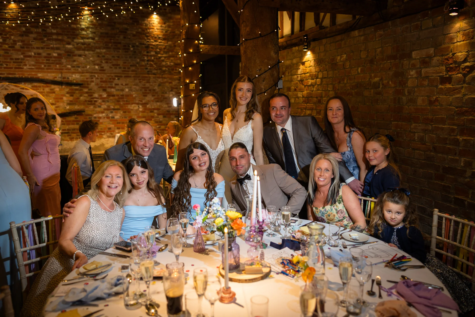 A large wedding party and family members smile together around a candlelit dining table inside a rustic brick barn venue draped in fairy lights.