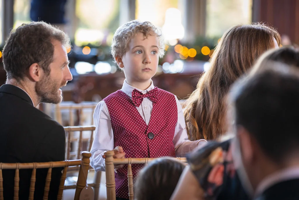 A young page boy wearing a red polka dot waistcoat and matching bow tie looks intently towards the front during a wedding ceremony.