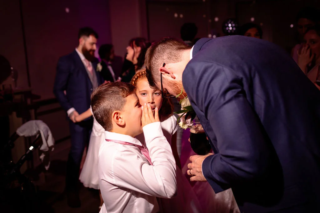 A young page boy whispers a secret to a flower girl while a guest in a navy suit leans in to listen during the evening reception.