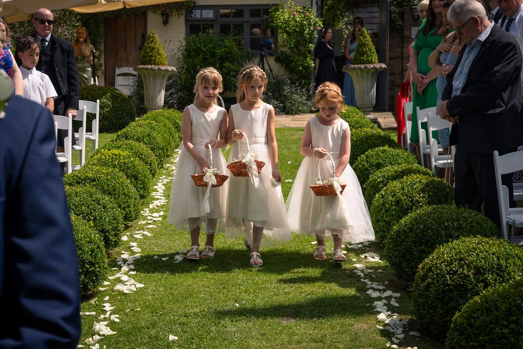 Three young flower girls walk down a grassy aisle lined with topiary bushes and scattered white petals during an outdoor wedding ceremony.