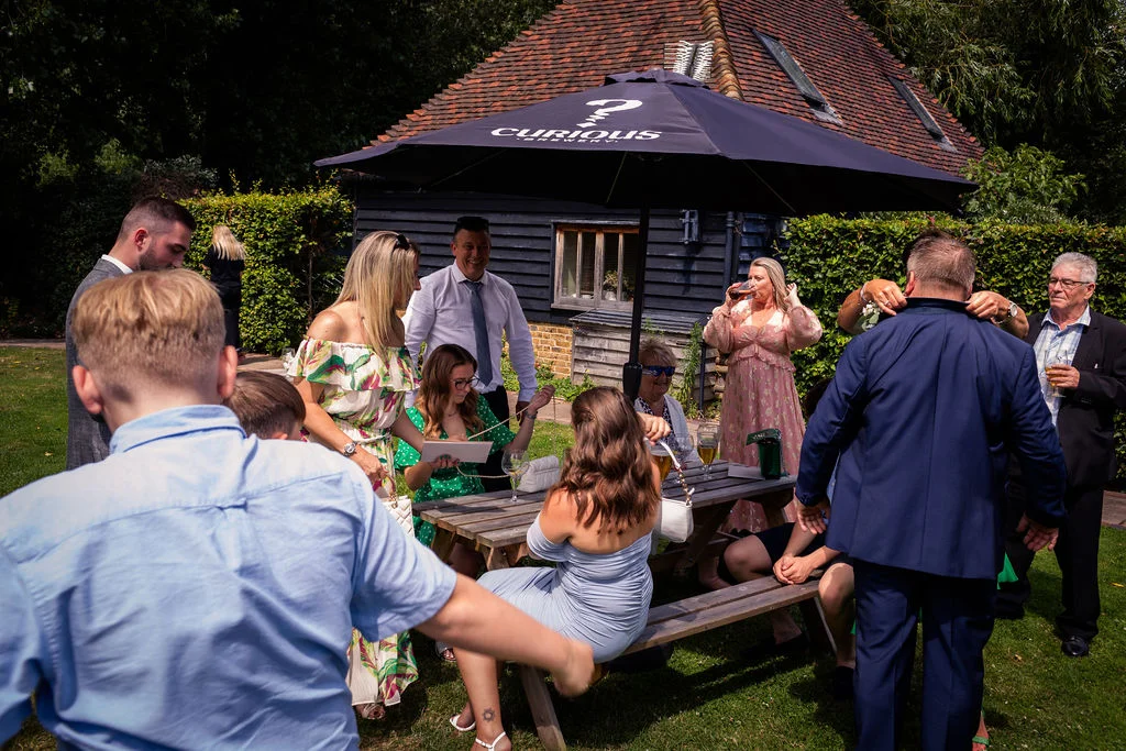 Wedding guests mingle and relax outside on a sunny day in the garden of a rustic barn venue with a branded black umbrella.