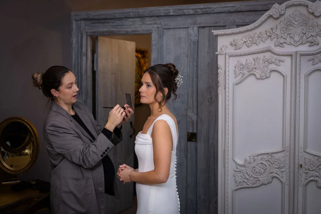 A makeup artist applies finishing touches to a bride wearing a structured white gown with a textured bun hairstyle adorned with baby's breath.