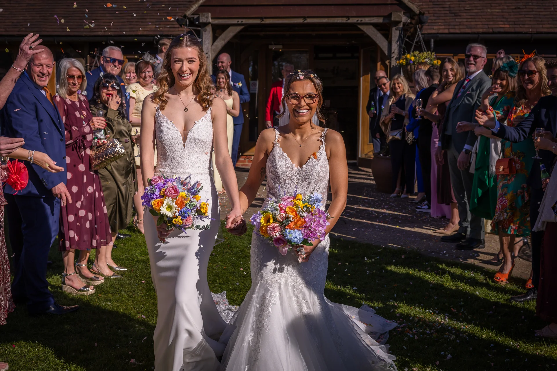 Two smiling brides walk hand in hand out of a rustic barn venue while joyful wedding guests shower them with colourful confetti in the bright sunshine.
