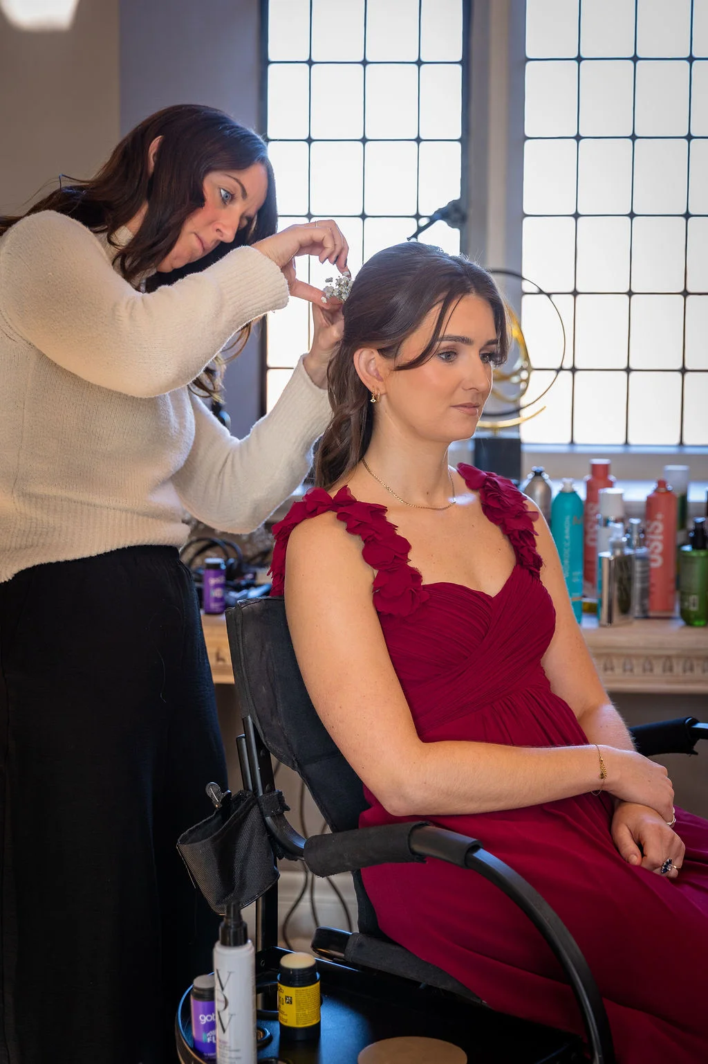 A hairstylist gently places small white flowers into the half-up hairstyle of a bridesmaid wearing a striking red dress during morning preparations.