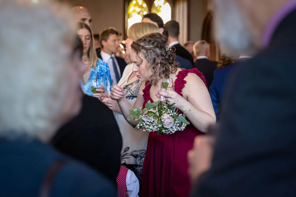 A bridesmaid in a burgundy dress holding a floral bouquet mingles with guests at a lively drinks reception.