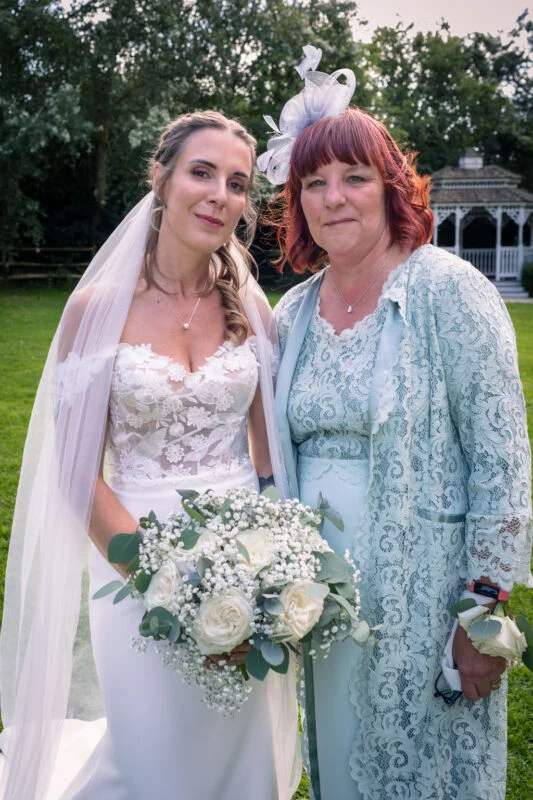 A smiling bride holding a white rose and baby's breath bouquet poses alongside her mother wearing a matching lace jacket and fascinator outdoors in a garden.