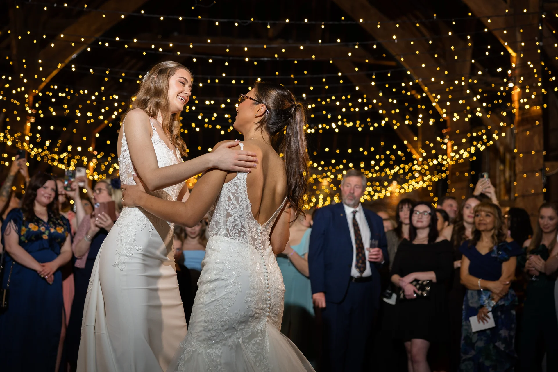 Two smiling brides share their first dance surrounded by a crowd of guests beneath a glowing canopy of fairy lights in a rustic barn venue.
