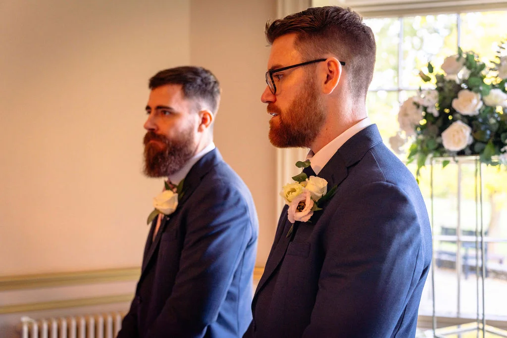Two grooms in sharp navy suits with floral buttonholes stand side by side during their indoor wedding ceremony.