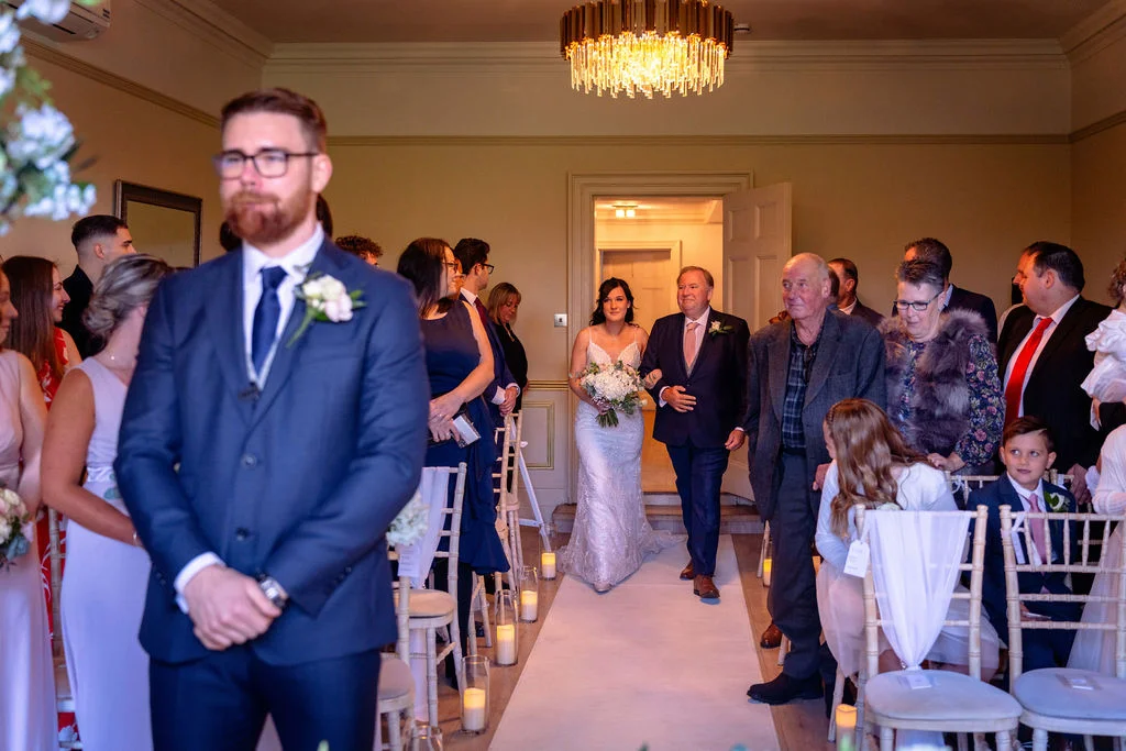 A groom in a navy suit waits at the altar while the bride walks down the aisle accompanied by her father during a civil ceremony.