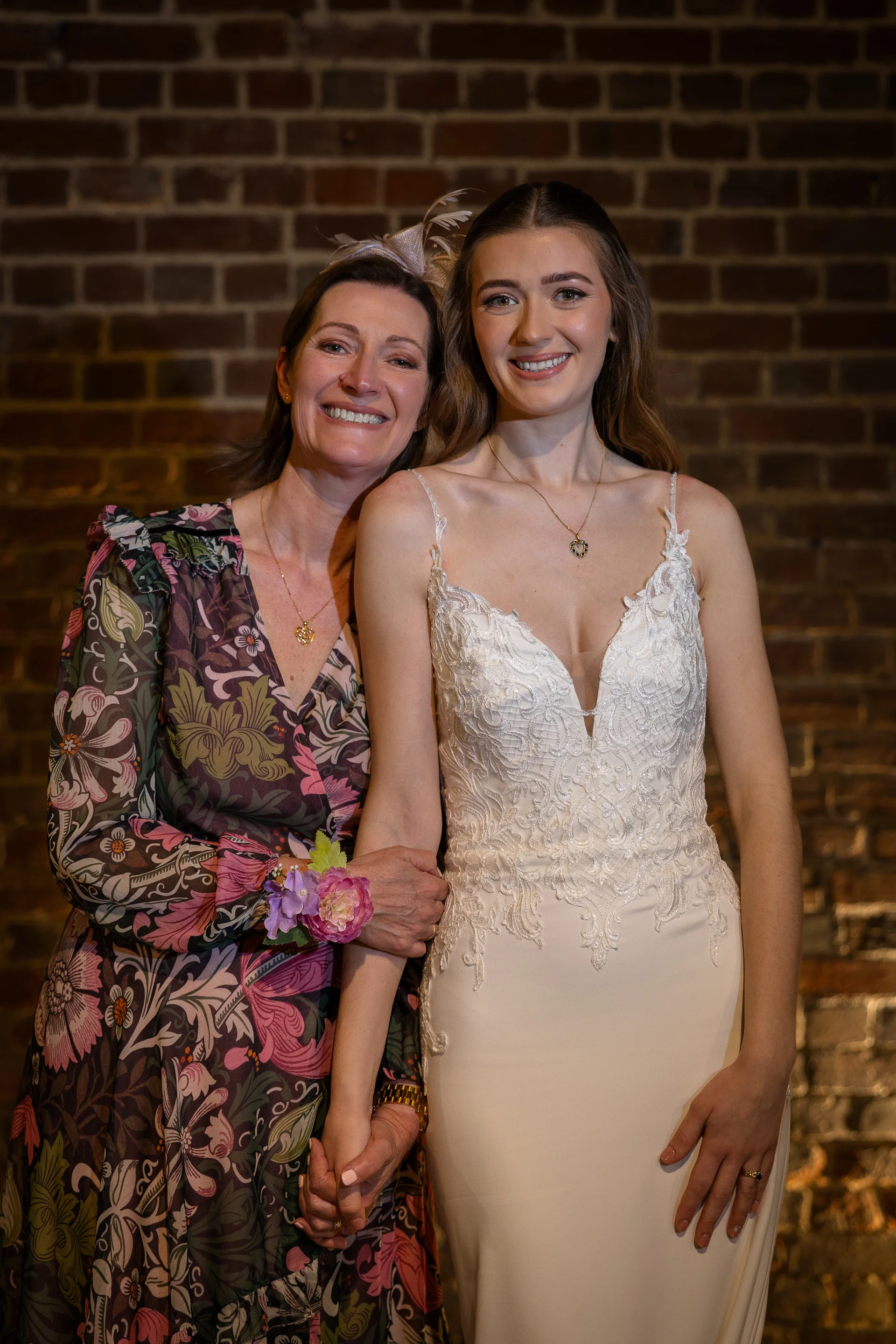 A smiling bride in a V-neckline lace wedding dress holding hands with her mother wearing a floral print dress against an exposed brick wall.
