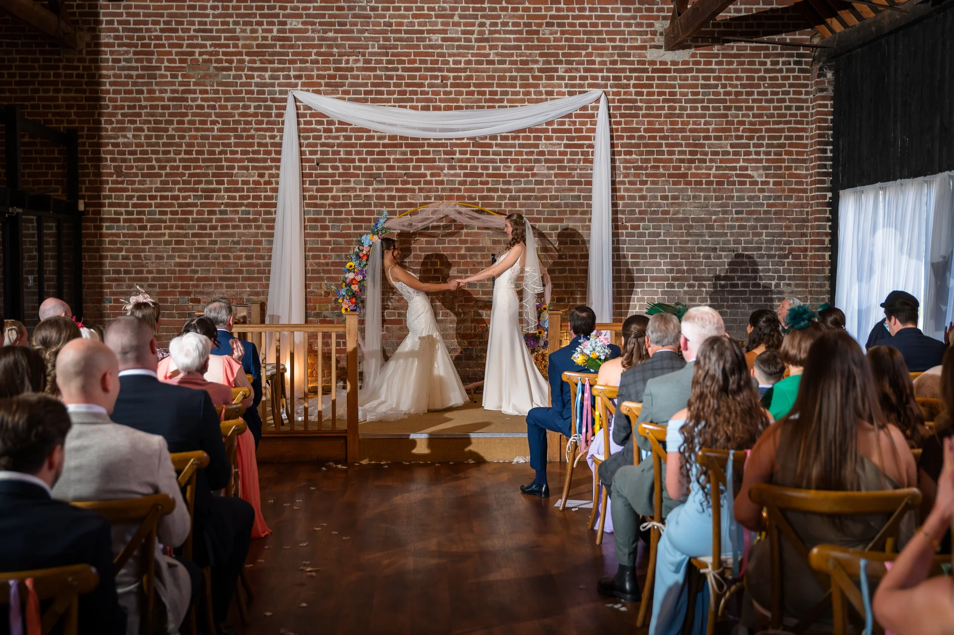 Two brides stand hand in hand during their wedding ceremony in a rustic brick barn, framed by a draped arch and seated guests.