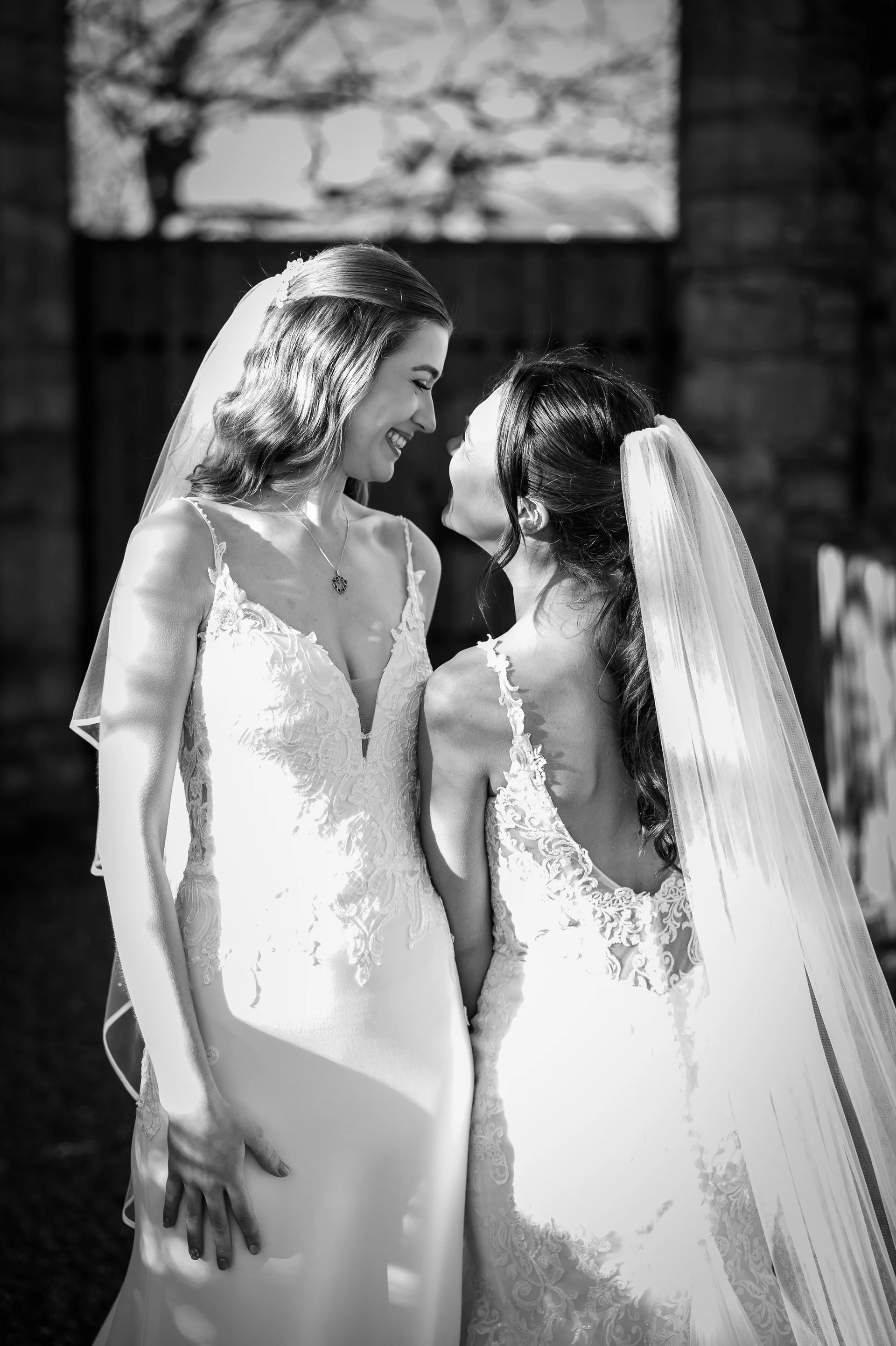 Two smiling brides in lace wedding gowns and veils stand face to face outdoors in a tender black and white portrait.