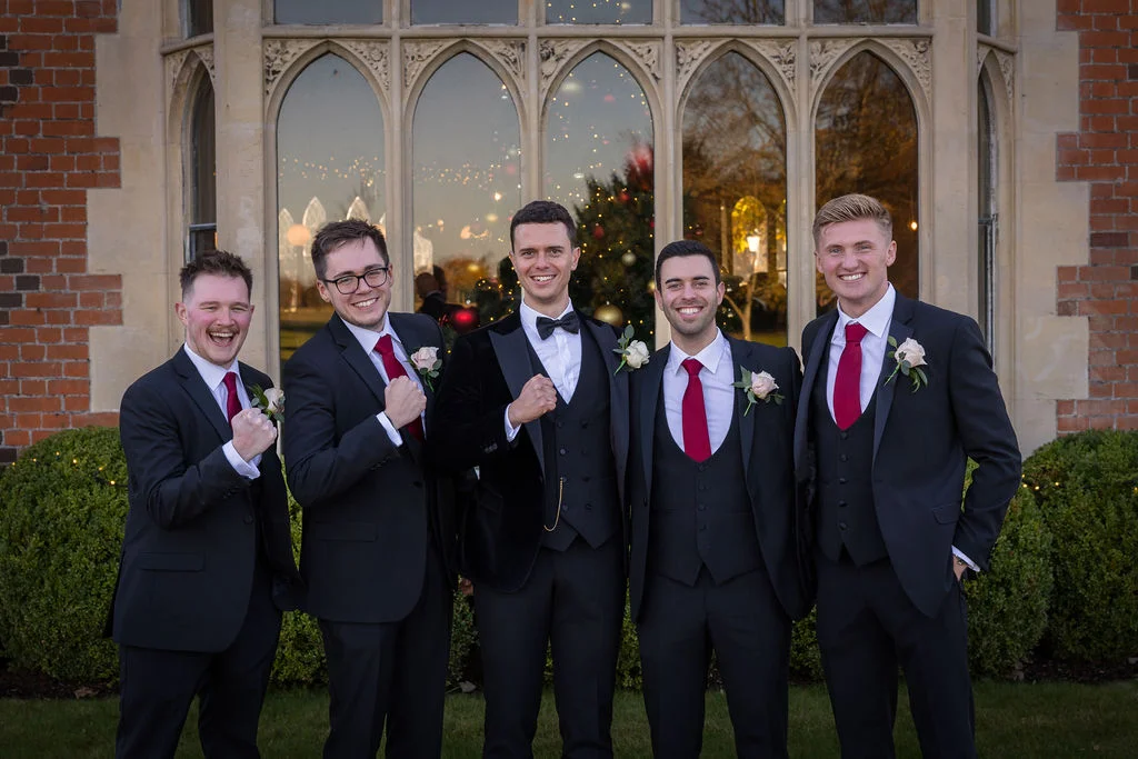 Five smiling groomsmen and the groom stand in front of a grand brick and stone building with large arched windows, wearing dark suits and matching red ties.