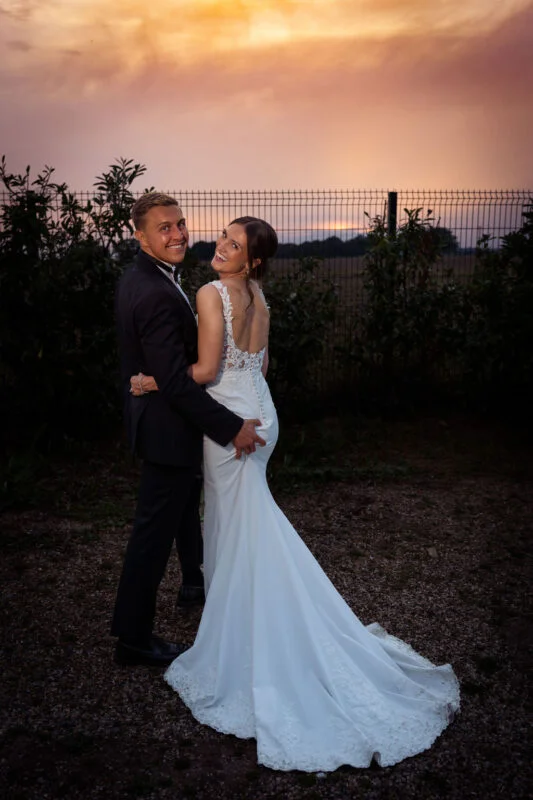 A bride and groom posing in front of a fence at sunset.