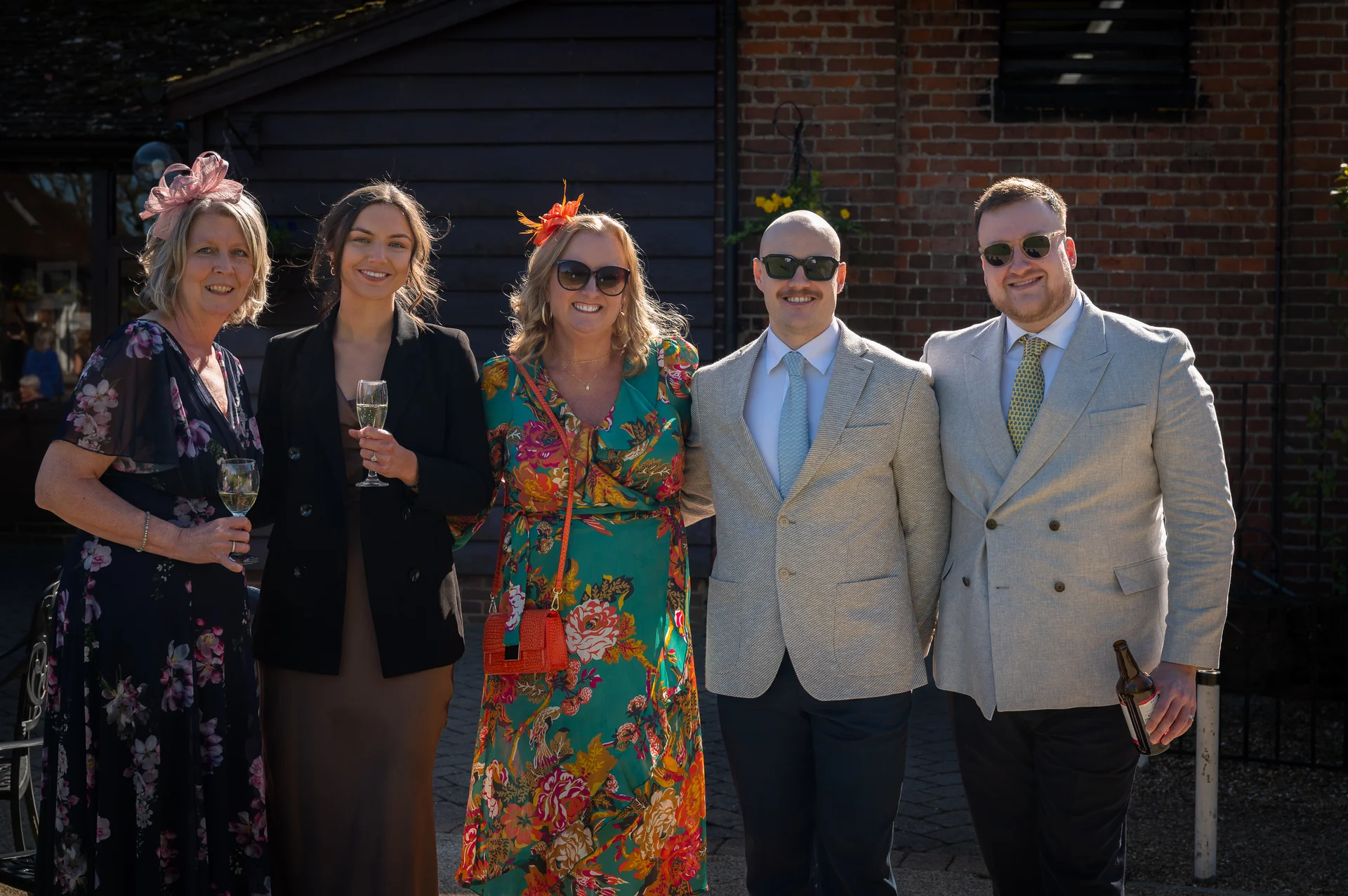 A group of five wedding guests dressed in formal attire and sunglasses pose together outdoors in the bright sunshine.
