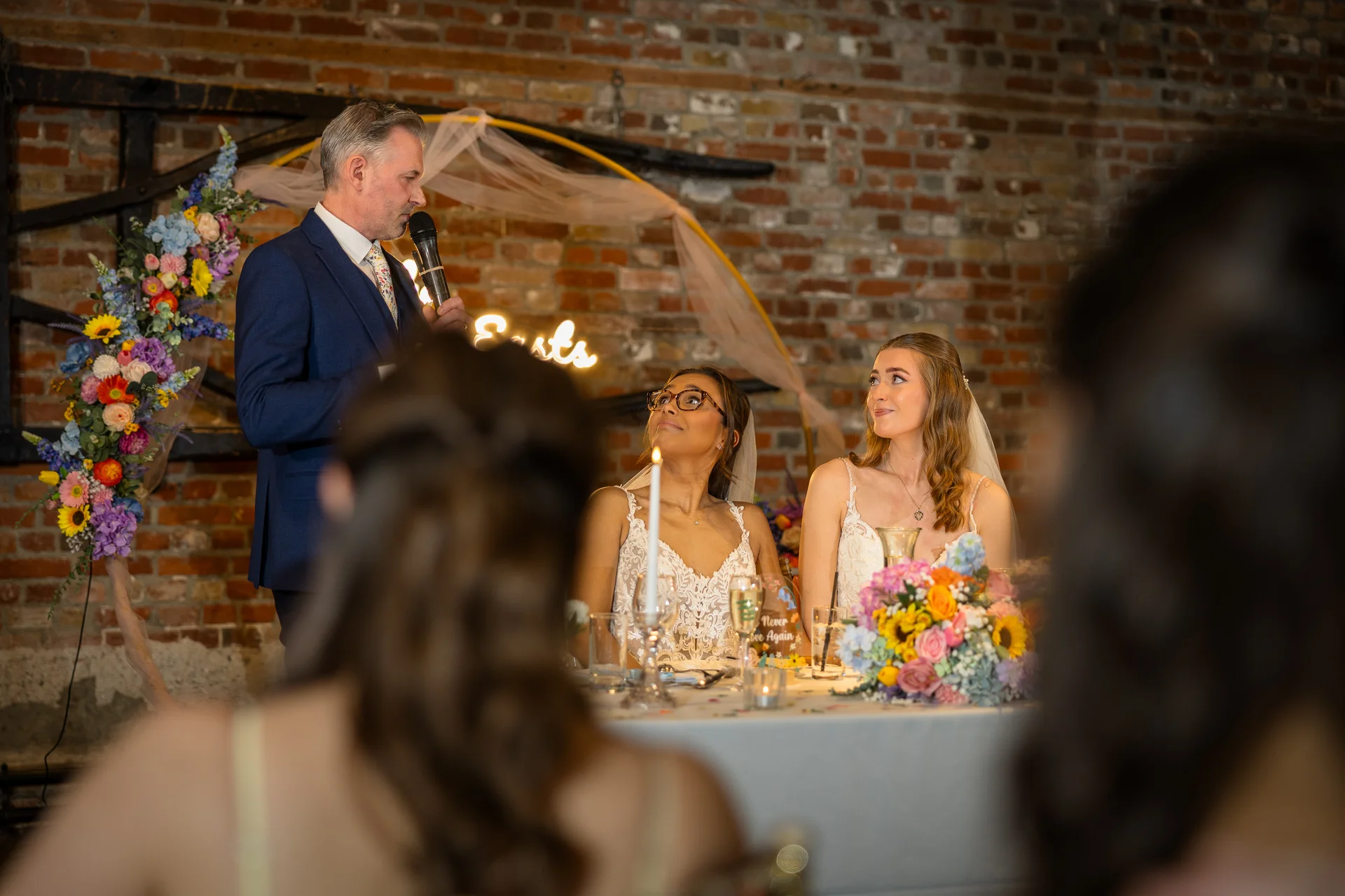A father gives a speech at the top table while two brides in white wedding dresses look up lovingly during their reception in a rustic brick barn.