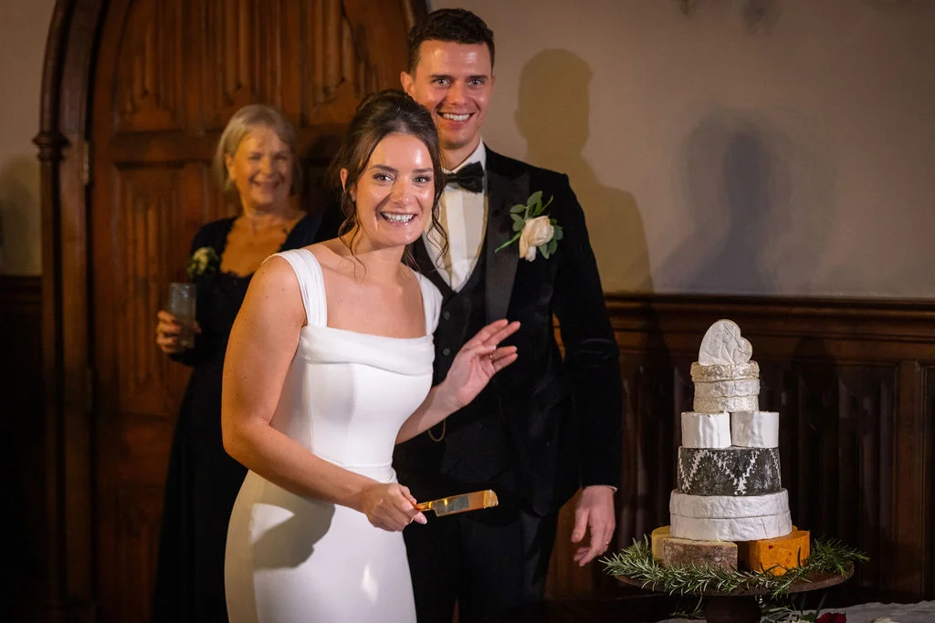 A smiling bride in a modern square-neck white gown prepares to cut a stacked cheese wheel wedding cake alongside her groom in a black tuxedo.