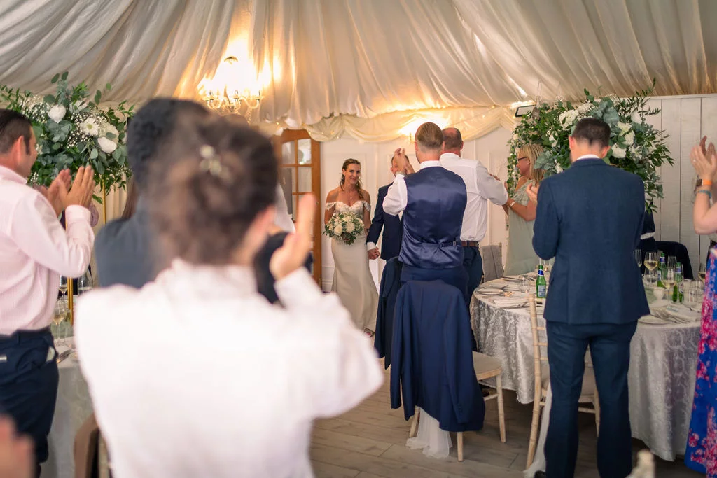 Newlyweds enter the marquee for the wedding breakfast as standing guests clap and cheer in celebration.