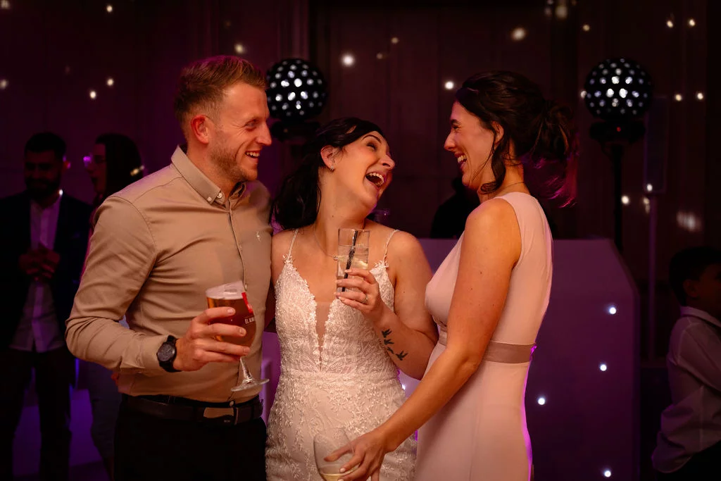 A bride in a white lace dress shares a joyous laugh with guests holding drinks during the evening reception.