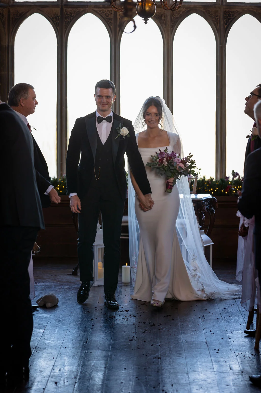 A newlywed couple walks hand in hand down the aisle of a historic venue with large arched windows and wooden flooring, smiling at their guests.