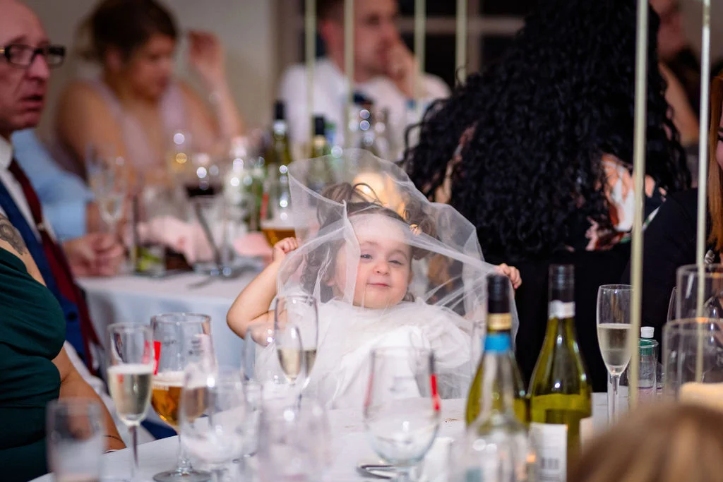A young flower girl playfully hides and peers through a sheer wedding veil while seated at a reception table during the wedding breakfast.
