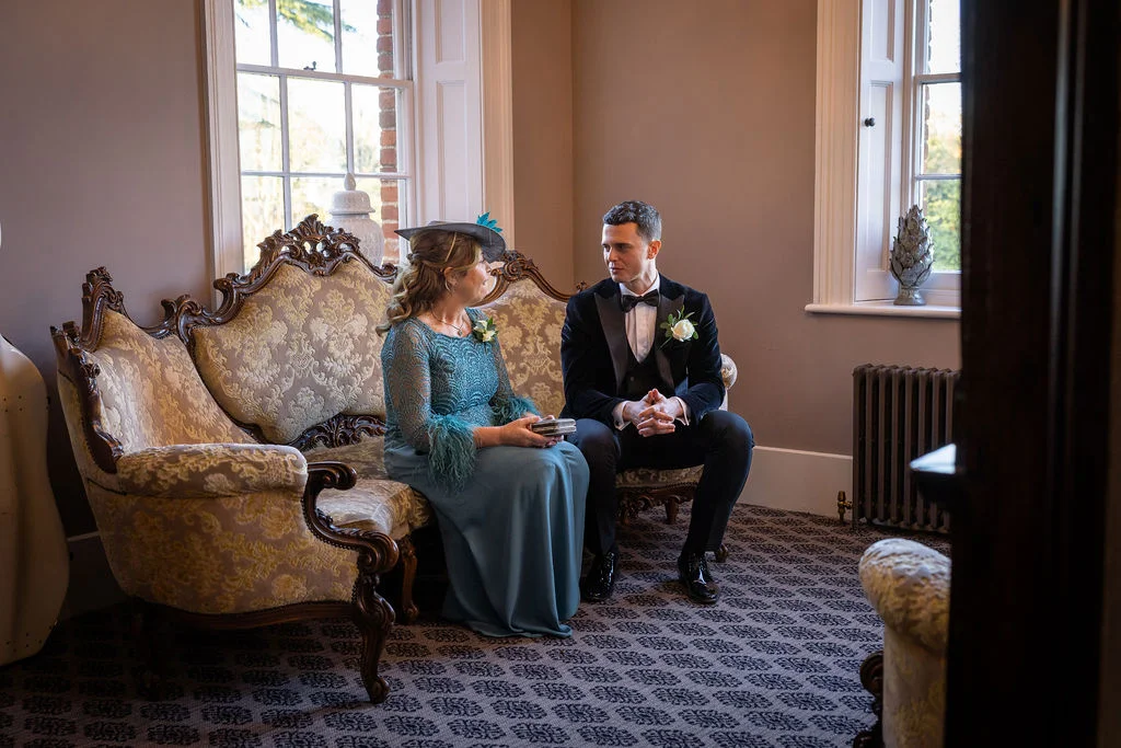 A mother and groom sit together on an ornate vintage sofa by a bright window inside a traditional country house venue.