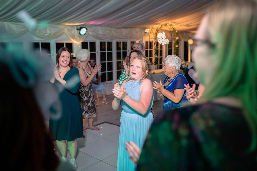 Wedding guests clap and cheer on the dance floor beneath a draped marquee ceiling during the evening reception.