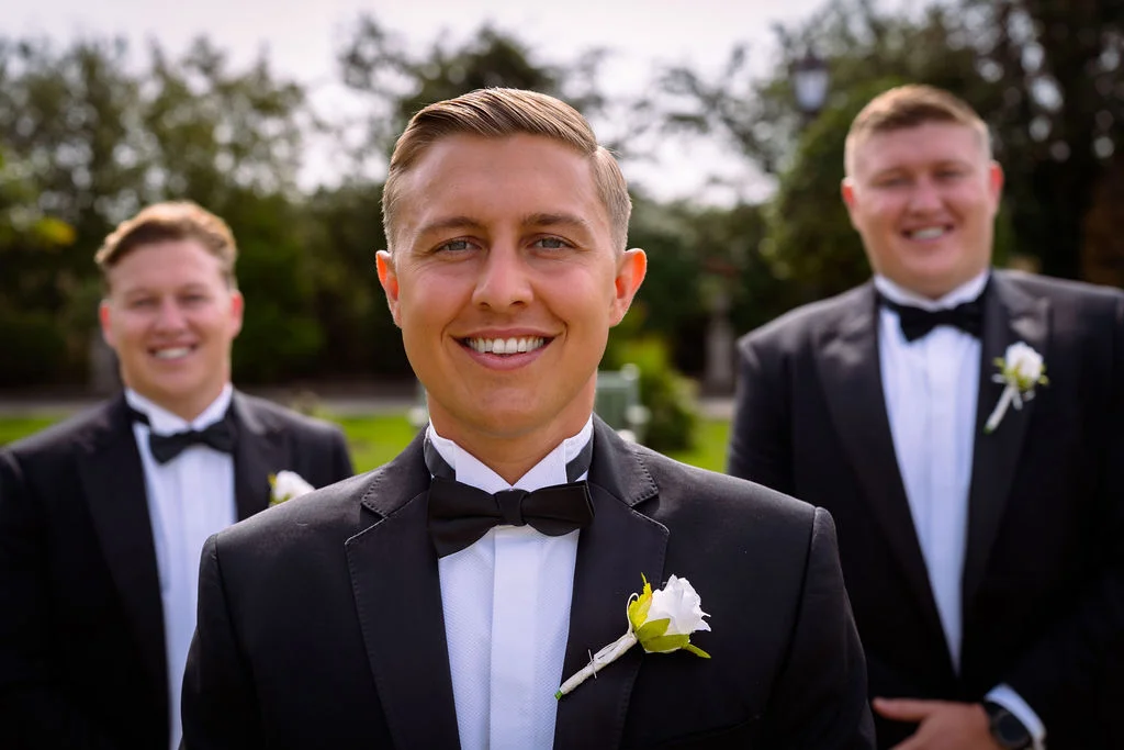Three smiling groomsmen wearing classic black tuxedos with bow ties and white rose boutonnieres pose together outdoors with a softly blurred garden background.