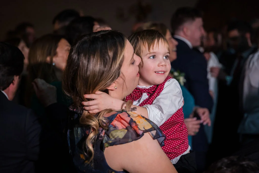 A guest holds a young child wearing a red polka-dot waistcoat during a wedding reception celebration.
