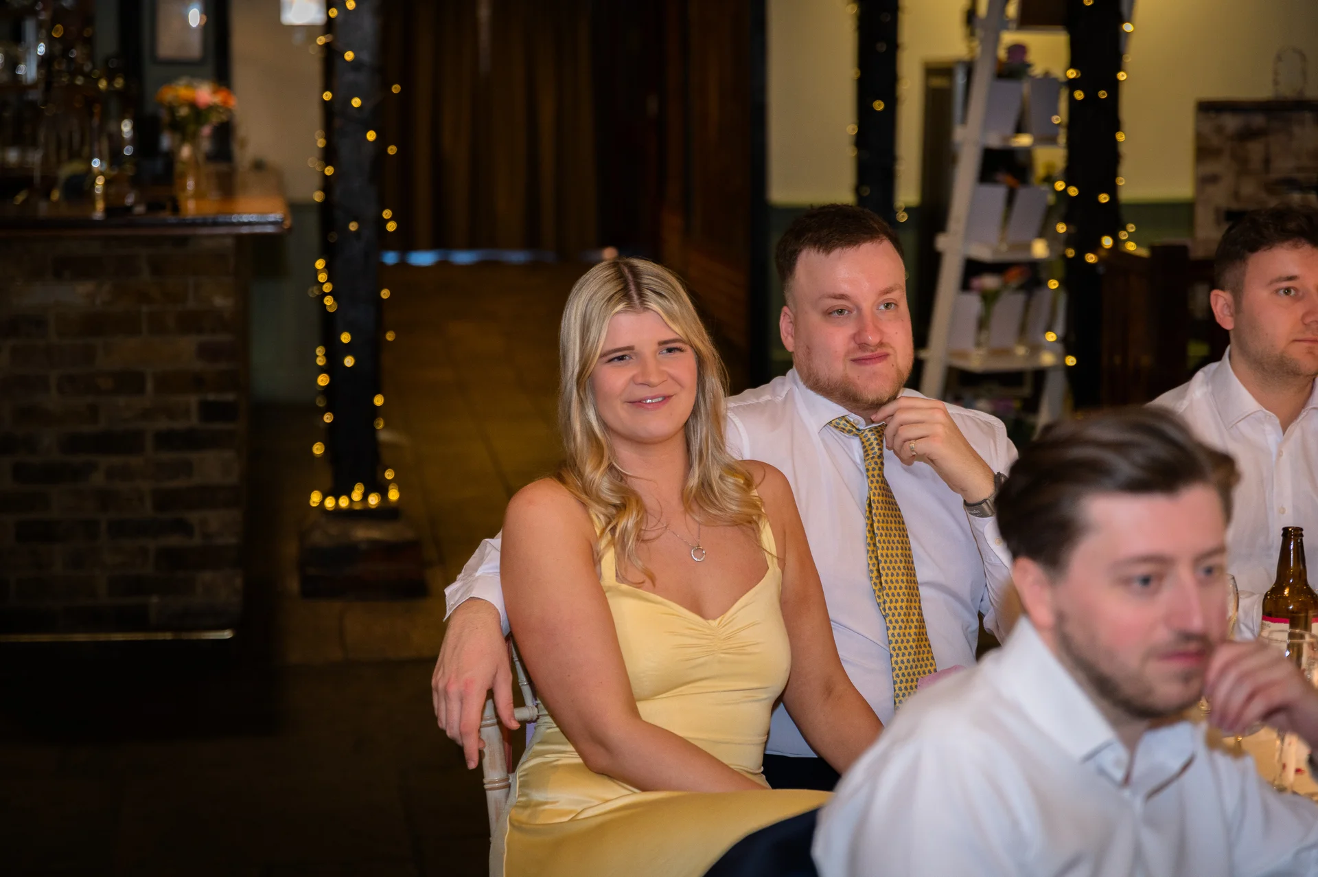 Wedding guests seated at tables during the reception speeches, smiling and listening attentively in a cosy venue illuminated by fairy lights.