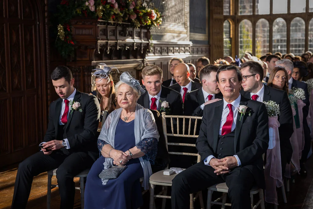 A mature woman wearing a navy dress and grey wrap sits alongside wedding guests in dark suits and red ties inside a wood panelled venue during a ceremony.