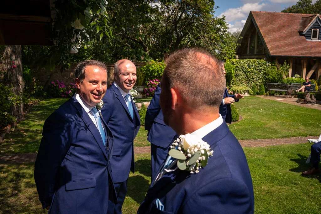Smiling groomsmen in navy suits gather in a sunlit manicured garden during a country house wedding.