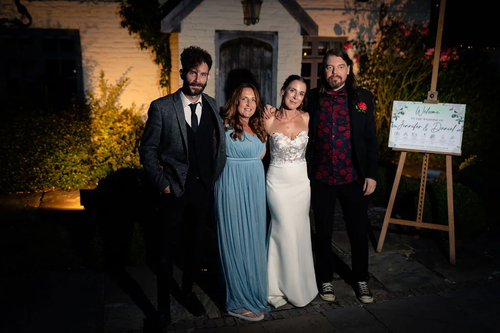 Four wedding guests and the bride pose together outside at night in front of a white building next to a welcome sign.