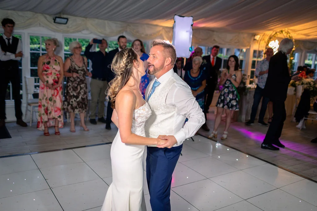 The newlyweds share a joyful first dance together on an illuminated dance floor inside a marquee surrounded by watching wedding guests.