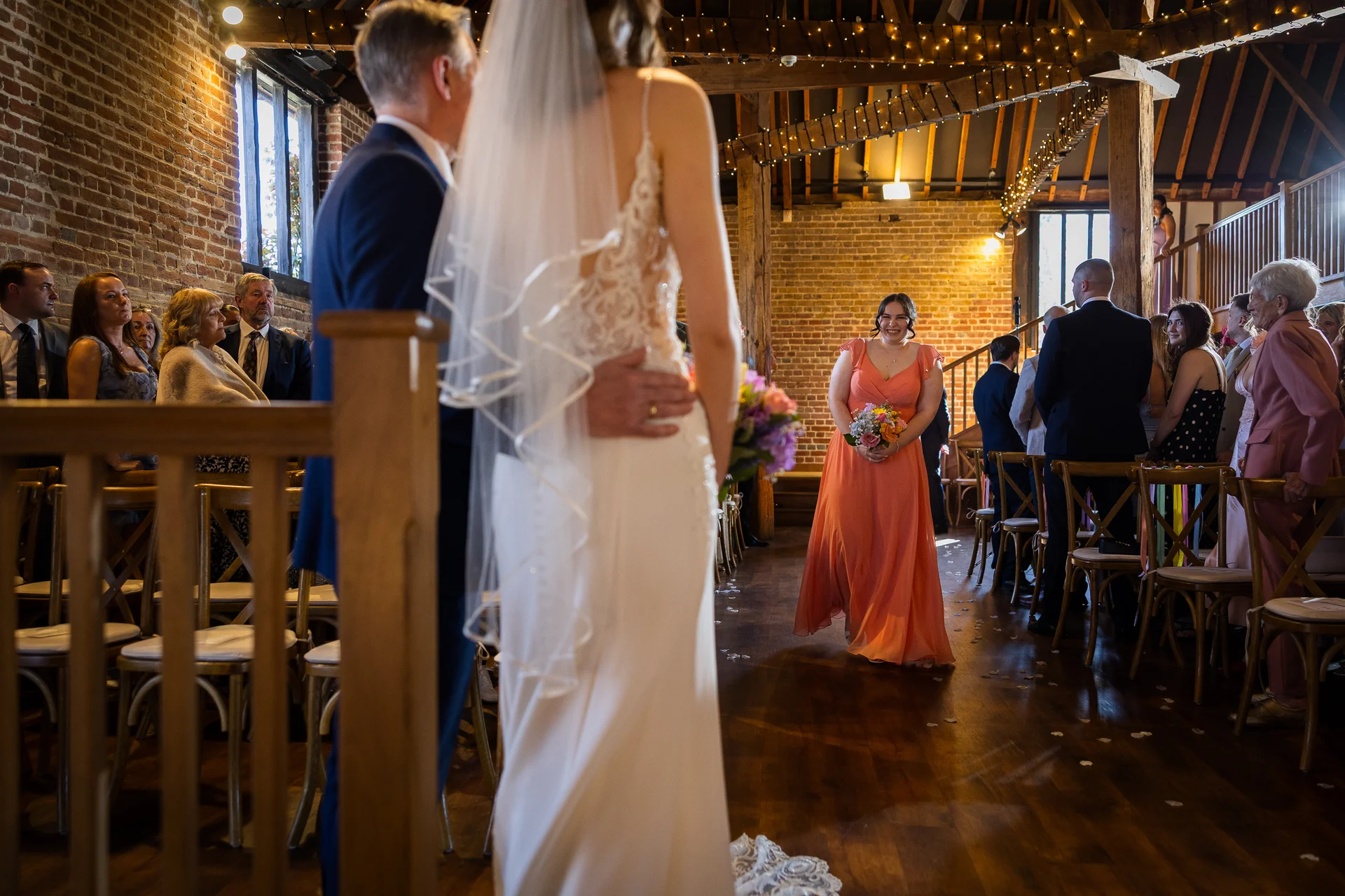 A bride in a lace-detailed gown and veil stands with the groom in a rustic brick barn, watching a bridesmaid in a flowing orange dress walk down the aisle carrying a bouquet.